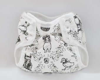 May include: A white cloth diaper with a black and white print of animals and flowers. The diaper has a white elastic waistband and white snaps.