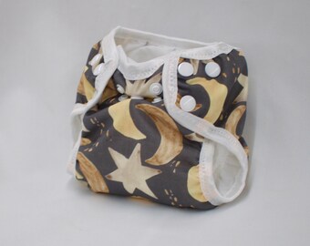 May include: A gray cloth diaper with a white trim and a pattern of yellow moons and stars. The diaper has snaps on the sides for adjusting the fit.
