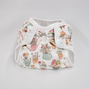 Op de afbeelding: Een witte katoenen luier met een bloemenprint van gieters en bloemen. De luier heeft witte drukknopen en een witte elastische tailleband.