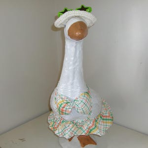May include: A whimsical goose figurine with a white body, brown beak, and feet. It wears a white crocheted hat with green star accents and a plaid dress with green, yellow, and red accents. The goose is standing on a white surface.