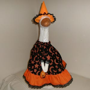 May include: A Halloween goose decoration wearing a witch costume. The costume includes a black dress with orange stars and crescent moons, an orange skirt, and a matching witch hat. The goose holds a small pumpkin.