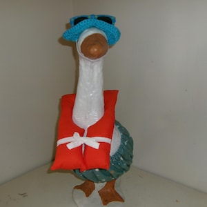 May include: A white goose figurine wearing a bright blue crocheted hat with black sunglasses on top, a vibrant orange life vest secured with a white ribbon tie, and a dark green skirt with a white polka dot pattern around its lower body.