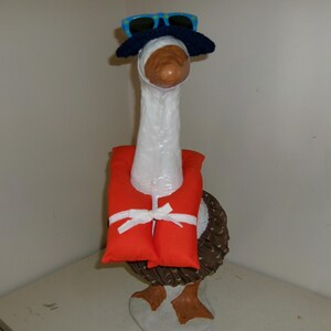 May include: A white goose figurine wearing a dark blue crocheted hat, blue and green patterned sunglasses, and a bright orange life vest secured with a white ribbon. The goose has brown feet and a brown textured base, standing in an upright position.