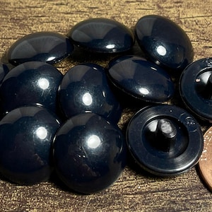 May include: A collection of navy blue buttons in various shapes, including oval and round, with a shiny, reflective surface. A US one-cent coin is included for size comparison. The buttons are scattered on a textured, brown surface.