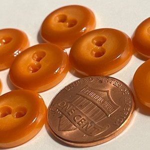 May include: Eight two-hole buttons in a light orange color. The buttons are arranged in a semi-circle with a penny in the center for scale.