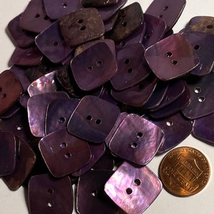 May include: A pile of purple iridescent shell buttons with two holes each. The buttons are square shaped with rounded corners. A US penny is included for scale.