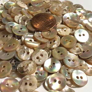 May include: A pile of approximately 100 two-hole iridescent mother of pearl buttons. The buttons are a pale cream color with a slight pink hue. The buttons are approximately 1/2 inch in diameter.