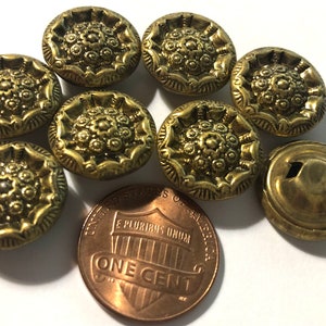 May include: A collection of antique brass buttons with a floral design. The buttons are round and have a detailed, raised pattern. A U.S. one-cent coin is included for size comparison. The buttons are likely for sewing or crafts.