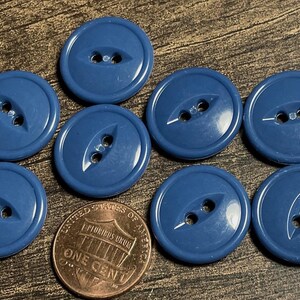 May include: A collection of eight navy blue buttons with two holes each, arranged on a wooden surface. A copper one-cent coin is included for size comparison. The buttons have a glossy finish and a slightly oval shape.