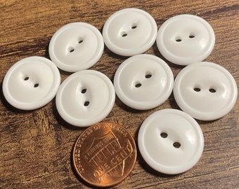 May include: Eight white plastic buttons with two holes each. The buttons are round and have a slightly raised edge. They are arranged in a circle around a penny.