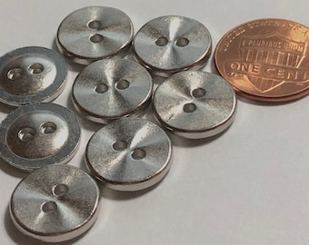 May include: Eight silver metal buttons with two holes each. The buttons are round and have a shiny, reflective surface. The buttons are arranged in a semi-circle around a penny.