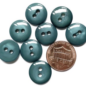 May include: A collection of teal-colored, round buttons with two holes each, scattered on a white surface. A copper one-cent coin is included for size comparison. The buttons have a glossy finish.