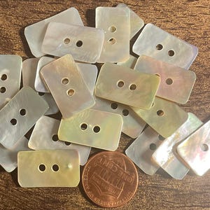 May include: A collection of rectangular, iridescent mother-of-pearl buttons with two holes each. The buttons are scattered on a wooden surface, with a US one-cent coin for size comparison. The buttons have a pearlescent sheen.