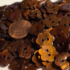 May include: A pile of brown, iridescent, flower-shaped buttons with two holes each. A US penny is included for scale.