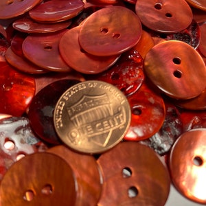 May include: A pile of red, iridescent, two-hole buttons. The buttons are oval shaped and have a shiny finish. A US penny is included for scale.