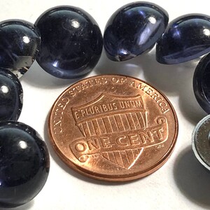 May include: A close-up of eight dark blue glass cabochons, each about the size of a US penny. The cabochons are arranged in a semi-circle around a penny, with one cabochon on top of the penny. The cabochons have a smooth, shiny surface.