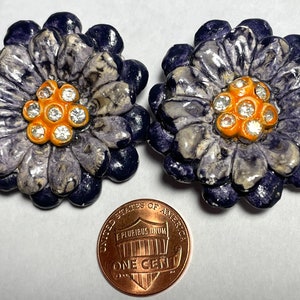 May include: A pair of vintage ceramic flower earrings with a purple and white marbled design. The center of each flower has a cluster of clear rhinestones set in an orange center.