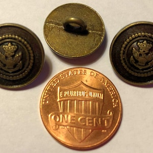 May include: Three antique brass buttons with a nautical design featuring an anchor, crown, and wreath. The buttons are dark brown with a gold-colored design. A penny is included for size comparison.