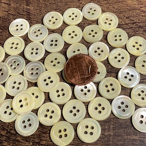 May include: A collection of round, off-white, four-hole buttons scattered across the frame. A single copper-colored one-cent coin is positioned among the buttons to provide a sense of scale for the small, circular fasteners.