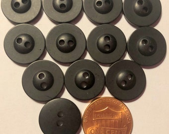 May include: A collection of 11 black plastic buttons with three holes each. The buttons are arranged in a circle with a penny in the center for size comparison.