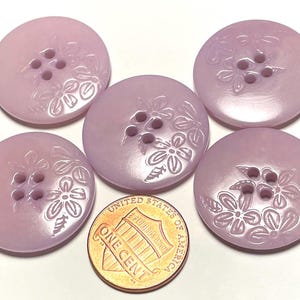 May include: Five round, light purple buttons with four holes each. The buttons have a floral design etched around the holes. A one-cent coin is next to the buttons for size comparison.