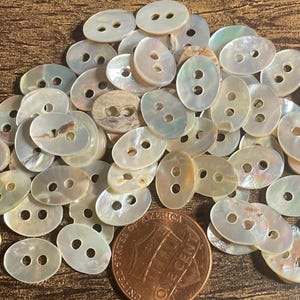 May include: A collection of oval-shaped, iridescent mother-of-pearl buttons with two holes each. A U.S. one-cent coin is included for size comparison. The buttons have a pearlescent sheen and vary slightly in color.