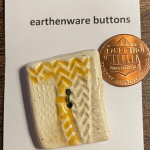 May include: A white ceramic button with a yellow zig-zag pattern and a green metal shank. The button is about the size of a US penny. The button is labeled "Logger's Daughter earthenware buttons" and "MACHINE WASHABLE".
