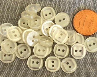 May include: A pile of 20 white plastic buttons with two holes each. The buttons are round with a slightly squared shape. The buttons are about the size of a UK penny.