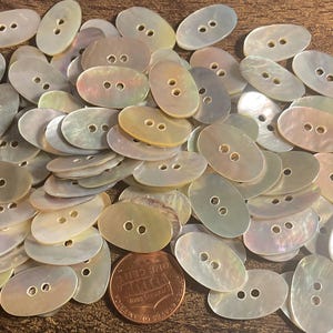 May include: A collection of iridescent oval-shaped buttons with two holes each. The buttons are in various shades of white, cream, and pale yellow. A penny is included for size comparison. These buttons are ideal for sewing and craft projects.
