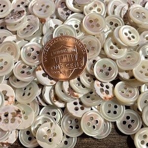 May include: A pile of white mother of pearl buttons with four holes each. The buttons are approximately the size of a US penny.