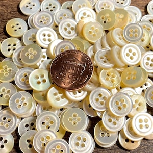 May include: A large pile of white and cream colored four-hole buttons. The buttons are made of a pearlescent material and are approximately the size of a US penny.
