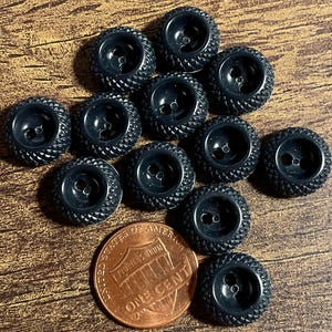 May include: A collection of twelve small, round, black buttons with a textured, cross-hatched rim and a smooth center featuring two holes. A copper-colored one-cent coin is positioned near the buttons for scale, measuring 0.75 inches in diameter.