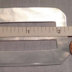 May include: A white, rectangular, plastic buckle with two rectangular cutouts. The buckle is approximately 1 inch wide and 2 inches long. A ruler and a penny are included for scale.
