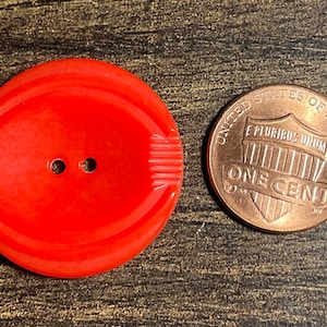 May include: A bright red, large button with two holes and a textured design, next to a United States one-cent coin. The button is round with a slightly oval shape.