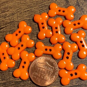 May include: A collection of bright orange, bone-shaped buttons with two holes each. A US one-cent coin is included for size comparison. The buttons are made of a shiny plastic material and are scattered on a wooden surface.