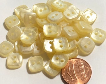 May include: A pile of 30 off-white, square-shaped buttons with two holes each. The buttons are slightly iridescent and have a pearly sheen. A penny is included for scale.