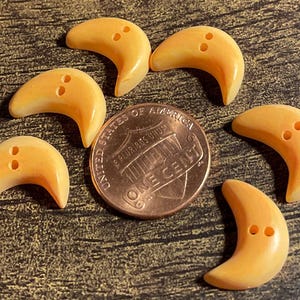 May include: Six crescent-shaped buttons in shades of yellow and orange, each with two holes. A penny is next to the buttons for size comparison. The buttons are on a textured, brown surface.