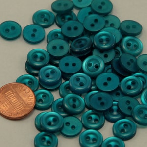 May include: A collection of teal-colored, two-hole buttons scattered on a surface. A copper penny is included for size comparison. The buttons are round and have a glossy finish, suitable for sewing or craft projects.