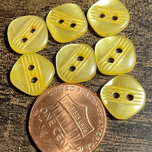 May include: A collection of eight pale yellow, square-shaped buttons with two holes and diagonal etched lines. A copper United States one-cent coin is included for size comparison. The buttons are likely for sewing or crafting.
