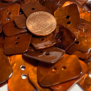 May include: A pile of brown, iridescent, two-hole buttons. The buttons are made of mother of pearl and have a shiny, glossy finish.