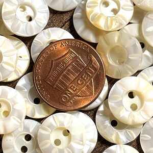 May include: A collection of white mother of pearl buttons with a raised floral design. The buttons have two holes and are arranged in a random pattern. A US penny is included for scale.