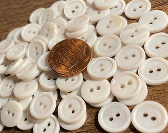 May include: A large pile of white, two-hole, round buttons. The buttons are made of a smooth, shiny material, possibly mother of pearl. A US penny is included for scale.