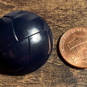 May include: A navy blue, spherical object with a textured surface, resembling a small ball, sits next to a copper one-cent coin. The coin displays the words "ONE CENT" and "UNITED STATES OF AMERICA".