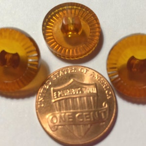 May include: Four amber-colored, round buttons with a ridged design and a central shank. A copper one-cent coin is next to the buttons for size comparison. The buttons have a translucent appearance.