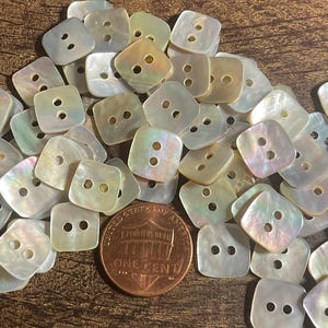 May include: A collection of iridescent, square-shaped buttons with two holes each. The buttons are scattered on a wooden surface, with a penny for size comparison. The buttons have a pearlescent sheen, reflecting various colors.