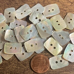 May include: A collection of iridescent, rectangular buttons with two holes each. The buttons are scattered on a wooden surface, with a US penny for size comparison. The buttons have a pearlescent sheen, reflecting various colors.