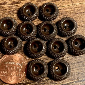 May include: A collection of eleven dark brown buttons, each featuring a textured, cross-hatched pattern on the outer rim and two central holes. A copper-colored United States one-cent coin is positioned next to the buttons for scale.