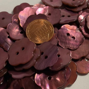 May include: A pile of purple iridescent shell buttons in a flower shape with two holes each. The buttons are arranged in a random pattern and a US penny is visible in the center of the pile.