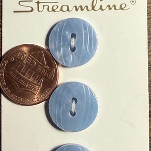 May include: Three round, light blue buttons with a subtle vertical striped pattern and two center holes, attached to a white display card labeled Streamline and Made in Thailand, shown next to a copper-colored one-cent coin for scale.