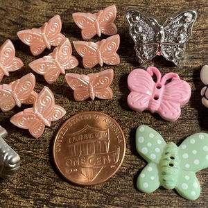 May include: A collection of butterfly-shaped embellishments in various colors and styles. Includes light pink, silver, and green butterflies, along with a penny for size comparison. Ideal for crafting and decorative projects.